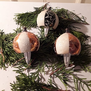 May include: Three vintage Christmas ornaments with a half-and-half design. One side is white with glitter, the other is gold or black with a starburst pattern. The ornaments are arranged on a bed of green evergreen branches.