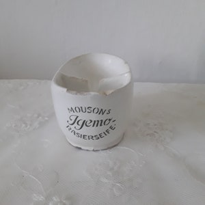 Könnte beinhalten: Ein weißer Rasierseifenhalter aus Keramik mit dem Text "MOUSONS Igemo RASIERSEIFE" in Schwarz. Der Halter hat eine gebogene Form mit einer gebrochenen Kante. Der Hintergrund ist eine weiße, strukturierte Oberfläche.