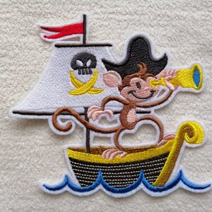 Könnte beinhalten: Gestickter Aufnäher mit einem Affenpiraten, der einen schwarzen Hut trägt und ein Teleskop hält, auf einem gelben und braunen Schiff mit Totenkopf und Knochen und einer Banane auf dem Segel segelt. Das Schiff segelt auf blauen Wellen.