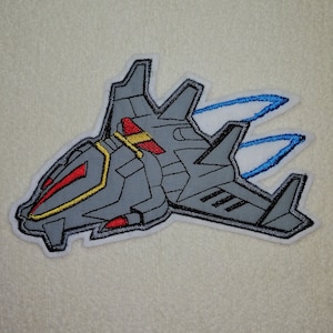 Könnte beinhalten: Gestickter Bügel-Patch mit einem grauen Raumschiff mit roten und gelben Akzenten, das mit blauen Flammen fliegt.