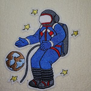 Könnte beinhalten: Ein gestickter Aufnäher mit einem Astronauten in einem blauen Raumanzug, der im Weltraum mit einem braunen und blauen Planeten und gelben Sternen schwebt.