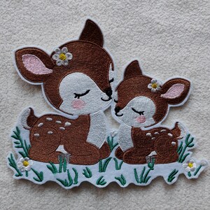 Könnte beinhalten: Gestickter Aufnäher mit zwei Hirschen, einem größeren und einem kleineren, in einer liebevollen Umarmung. Die Hirsche sind braun mit weißen und rosa Akzenten und kleinen Blumendetails. Sie sind vor einem Hintergrund aus grünem Gras und weißen Blumen platziert.
