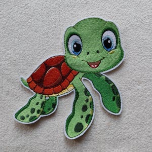 Könnte beinhalten: Gestickter Aufnäher einer Cartoon-Meeresschildkröte. Die Schildkröte hat einen grünen Kopf und Gliedmaßen mit einem rot-orangen Panzer. Die Augen der Schildkröte sind blau und sie hat einen lächelnden Mund. Der Aufnäher hat einen weißen Rand.