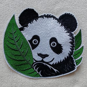 Könnte beinhalten: Gestickter Aufnäher mit einem Panda, der hinter einem grünen Blatt hervorlugt. Der Panda hat schwarz-weißes Fell mit gestickten Details. Das Blatt ist leuchtend grün, und der Aufnäher befindet sich auf hellem Hintergrund.
