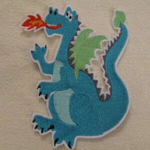 Könnte beinhalten: Ein blauer gestickter Drache mit grünen Flügeln und einer roten Flamme, die aus seinem Maul kommt. Der Drache steht auf seinen Hinterbeinen mit seinem Schwanz um seinen Körper gewickelt.