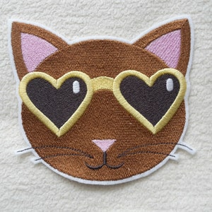 Könnte beinhalten: Gesticktes braunes Katzen Gesicht mit rosa Ohren und gelben herzförmigen Sonnenbrillen.