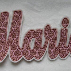 Könnte beinhalten: Rosa Stoffapplikation mit dem Namen "Marie" in einer kursivschrift. Der Stoff hat ein braunes und rosa Muster.