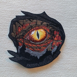 Könnte beinhalten: Gestickter Aufnäher mit einer Nahaufnahme eines Drachenauges in einem rot-orange-gelben Farbschema. Das Auge ist von einem schwarzen, strukturierten Hintergrund umgeben.