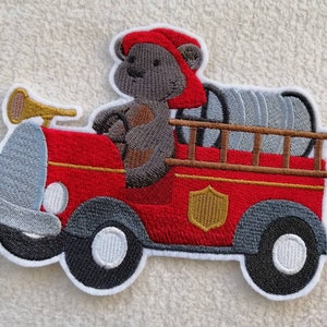Könnte beinhalten: Gestickter Bügel-Patch mit einem roten Feuerwehrauto, in dem ein grauer Bär mit roter Mütze sitzt und fährt. Das Auto hat einen Schlauch hinten und ein goldenes Schild an der Seite.