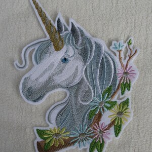 Könnte beinhalten: Gestickter Bügel-Patch mit einem weißen Einhornkopf, einem goldenen Horn und einem blauen Auge. Das Einhorn ist von grünen Blättern und rosa, blauen und gelben Blumen umgeben.