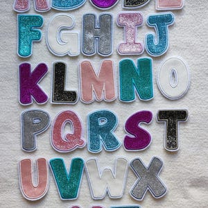 Könnte beinhalten: Eine Sammlung von sechsundzwanzig einzelnen Buchstaben-Aufnähern, die in Reihen angeordnet sind. Jeder Buchstabe weist eine ausgeprägte glitzernde oder einfarbige Textur in verschiedenen Farben wie Pink, Blau, Lila, Schwarz, Silber und Weiß auf.