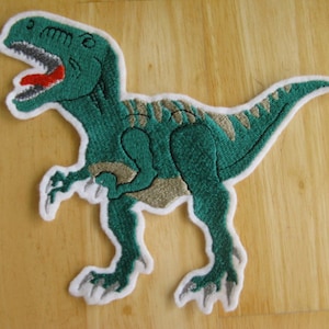 Könnte beinhalten: Gestickter Aufnäher mit einem grünen Dinosaurier und weißem Rand. Der Dinosaurier hat ein detailliertes Design mit roter Zunge, grauen Zähnen und beigen Akzenten. Geeignet für Kleidung oder Accessoires.