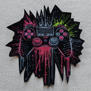 Könnte beinhalten: Schwarzer, gestickter Aufnäher mit einem Videospiel-Controller-Design. Der Controller ist in Pink und Blau umrandet, mit pinken und grünen Farbspritzern. Der Aufnäher hat einen gezackten, sternförmigen Rand.