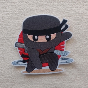 Könnte beinhalten: Gestickter Aufnäher mit einem Cartoon-Ninja in geduckter Pose. Der Ninja ist in Schwarz mit einem roten Schärpenband und einem Schwert gekleidet. Im Hintergrund befindet sich eine rote Sonne, und der Aufnäher hat einen hellblauen Rand.