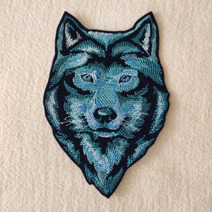 May include: Embroidered patch of a wolf's head in blue and black. The wolf's fur is a light blue with a darker blue outline. The wolf's eyes are black with a white highlight. The wolf's nose is black. The wolf's mouth is closed and the wolf's teeth are not visible.