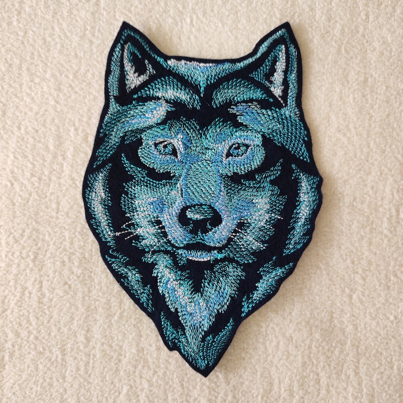 May include: Embroidered patch of a wolf's head in blue and black. The wolf's fur is a light blue with a darker blue outline. The wolf's eyes are black with a white highlight. The wolf's nose is black. The wolf's mouth is closed and the wolf's teeth are not visible.
