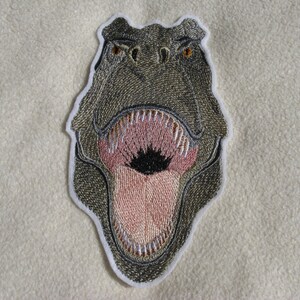 Könnte beinhalten: Gestickter Aufnäher mit dem Kopf eines Dinosauriers mit offenem Maul, das scharfe Zähne zeigt. Die Augen des Dinosauriers sind braun und der Mund ist rosa.