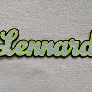 Könnte beinhalten: Ein weißer Filz-Patch mit schwarzen und neongrünen Umrandungen, auf dem der Name "Lennard" steht.