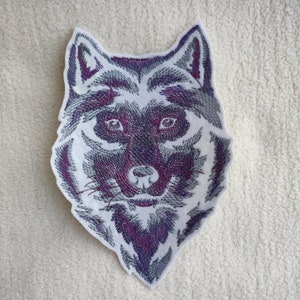 May include: Embroidered wolf head patch with a white background and purple and gray thread. The wolf's eyes are black and the nose is pink.