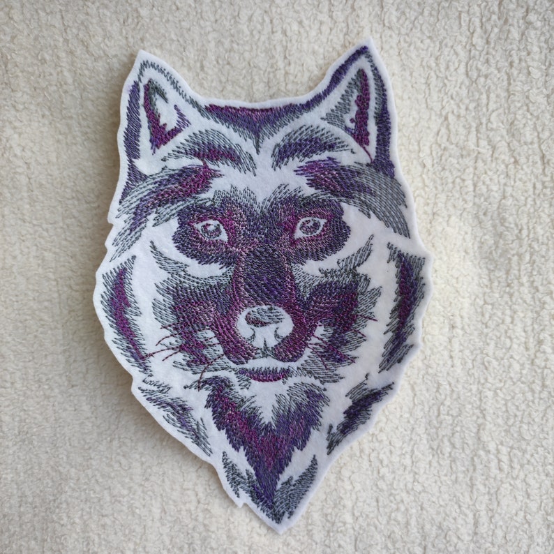 May include: Embroidered wolf head patch with a white background and purple and gray thread. The wolf's eyes are black and the nose is pink.