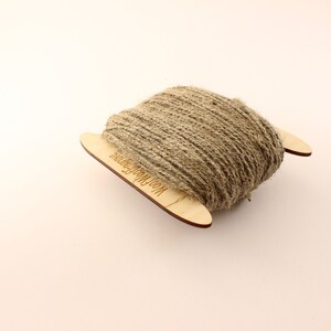 May include: A spool of natural twine wrapped around two wooden dowels. The dowels are labeled "Wool & More Fibers".