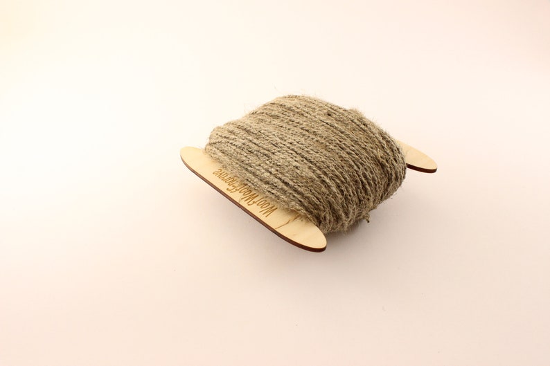 May include: A spool of natural twine wrapped around two wooden dowels. The dowels are labeled "Wool & More Fibers".