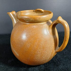 May include: A round, golden-brown ceramic teapot with a smooth, matte finish. The teapot features a wide, open top rim, a curved spout on the left side, and a sturdy, arched handle on the right side, showcasing a consistent, earthy color tone.