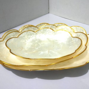 Puede incluir: Juego de tres bandejas decorativas en forma de nube. Las bandejas tienen un interior blanco nacarado y un borde dorado. Las bandejas están anidadas, con la más grande en la parte inferior y la más pequeña en la parte superior. Las bandejas son de material brillante.