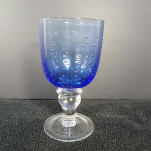 May include: A blue wine glass with a clear stem and base. The bowl of the glass is a vibrant blue with small, bubble-like inclusions. The stem is clear and bulbous, and the base is a flat circle.
