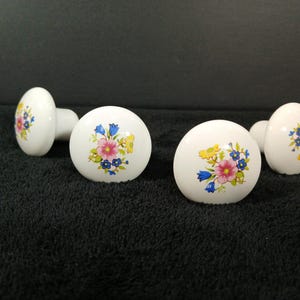 May include: Four white ceramic cabinet knobs with a rounded top surface. Each knob features a central floral decal with pink, blue, and yellow flowers surrounded by green leaves. The knobs have a smooth, glossy finish and a cylindrical base.