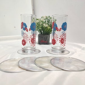 以下が含まれることがあります： 赤と青の花柄が施された2つの透明なグラス。それぞれ透明なコースターの上に置かれています。4つの追加のコースターは、真珠光沢のある仕上がりで手前にあります。小さな鉢植えの植物が背景にあります。