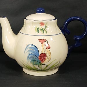 May include: Cream-colored ceramic teapot with a blue handle and accents. The teapot features a hand-painted rooster design on the front, with a blue tail, red comb, and yellow body. The lid is white with a blue knob.