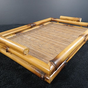 May include: A rectangular bamboo tray with a woven center. The tray features a natural bamboo frame with darker brown accents at the joints. A small, rounded bamboo handle is on one side. The tray is designed for serving or display.