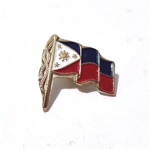 May include: A gold-coloured pin badge in the shape of the Philippine flag. The flag design includes a white triangle with a sun and stars, a blue stripe, and a red stripe. The badge has a gold border and a decorative element on the left.