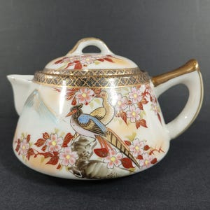 May include: A white ceramic teapot with a lid, featuring a hand-painted design of a bird, cherry blossoms, and a mountain scene. The handle and spout are white, with gold accents. The teapot is a decorative item.