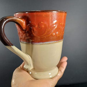 May include: A ceramic mug featuring a two-tone glaze with a glossy burnt orange upper section decorated with embossed scrollwork and a smooth cream-colored lower section. The mug has a curved handle that transitions from dark brown to cream.