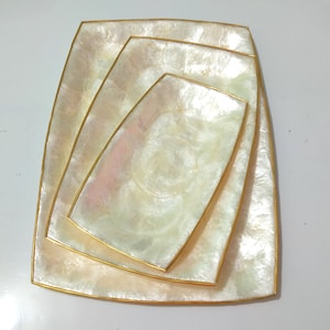 Puede incluir: Un juego de cuatro bandejas decorativas anidadas. Las bandejas tienen una superficie nacarada blanquecina con un borde dorado. Las bandejas son cuadradas con esquinas redondeadas, y cada una es de un tamaño ligeramente diferente.