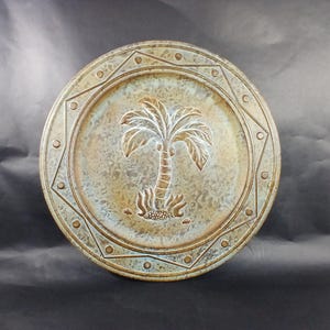 May include: Round decorative plate with a palm tree design. The plate features a raised palm tree in the center, surrounded by a geometric border with small circular accents. The color palette includes shades of brown, green, and gold.