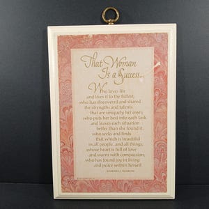 May include: A framed wall hanging with a cream-colored border and a decorative pink and white patterned background. The text reads "That Woman Is a Success..." followed by a poem. A gold-colored ring is attached at the top for hanging.