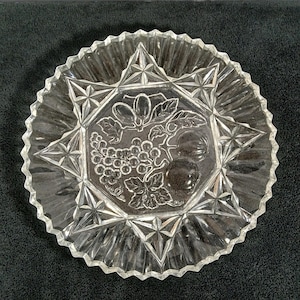 May include: A clear pressed glass plate featuring a central hexagonal motif decorated with embossed grapes, leaves, and fruit. The plate has a scalloped, serrated edge and a geometric star pattern radiating from the center to the outer rim.