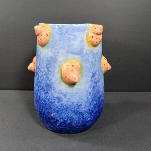 May include: A handmade ceramic vase with a blue gradient finish. The vase features several raised, rounded, orange-colored accents with small red dots. The vase has a unique, textured appearance and is suitable for home decor.