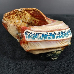 May include: A ceramic bowl shaped like a seashell with a glossy finish. The interior features a mottled brown and cream pattern. The exterior has a cream-colored band inlaid with small, angular, bright blue glass pieces against a textured surface.