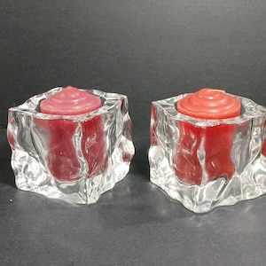 May include: Two clear glass candle holders with a textured, faceted design. Each holder contains a red candle. The candles have a circular, layered design on top. The candle holders are on a dark surface.