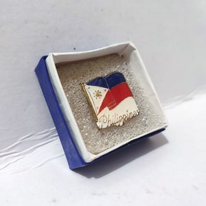 May include: A rectangular pin featuring the flag of the Philippines, with blue, red, white, and gold colors. The pin is in a blue and white box with the word "Philippines" in script. The pin is likely made of metal.