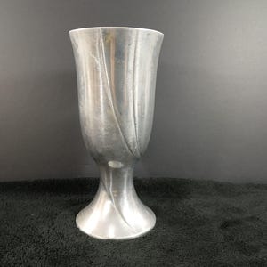 May include: A silver-colored vase with a flared top and a decorative, curved design. The vase has a pedestal base and a smooth, metallic finish. The background is dark.