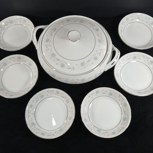 May include: A white porcelain serving bowl with a lid and handles, surrounded by six matching small bowls. Each piece features a delicate floral pattern in light blue and gray, with a silver trim. The set is designed for serving and dining.