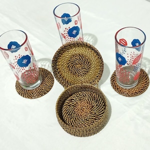 May include: Four tall clear drinking glasses with blue floral designs and red accents, resting on woven rattan coasters. A set of matching rattan coasters and a small basket are also present, creating a cohesive drinkware set.