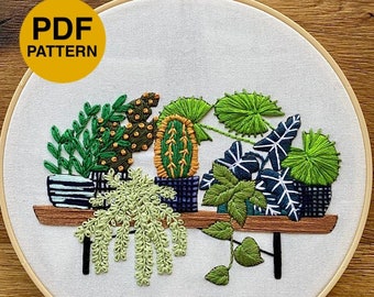 Può includere: Un telaio da ricamo contenente un pannello di tessuto bianco con un disegno ricamato di varie piante in vaso su una mensola di legno. Le piante includono cactus, succulente e fogliame in tonalità di verde, giallo e blu scuro. È visibile un cerchio giallo con il testo PDF PATTERN.