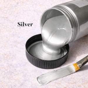 Silver metallic acrylic paint pouring from a jar into a lid with a matching silver brushstroke, shown in a square product image on a soft marble background.