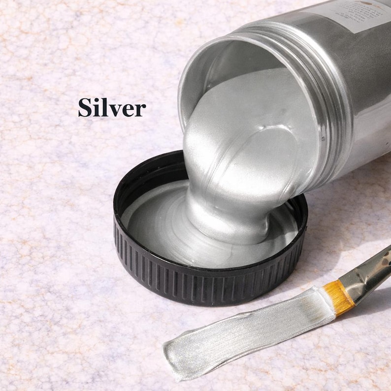 Silver metallic acrylic paint pouring from a jar into a lid with a matching silver brushstroke, shown in a square product image on a soft marble background.